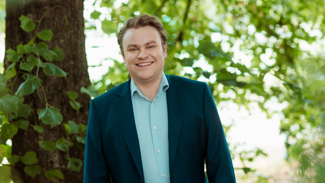 Daniel Naper med blondt hår iført en mørk dress og lys skjorte står med armene ned langs siden og smiler mot kameraet. Foto
